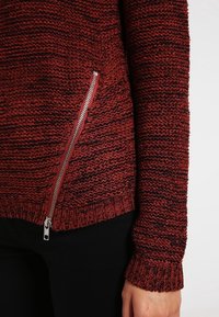 Pull en tricot couleur rouge foncé avec motif texturé, présentant une fermeture éclair argentée sur le côté et des poignets côtelés. Fabriqué en matériau en fil doux.