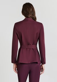Blazer su misura bordeaux con vita a cintura, silhouette allungata e texture liscia. Presenta una fessura sul retro e maniche lunghe.