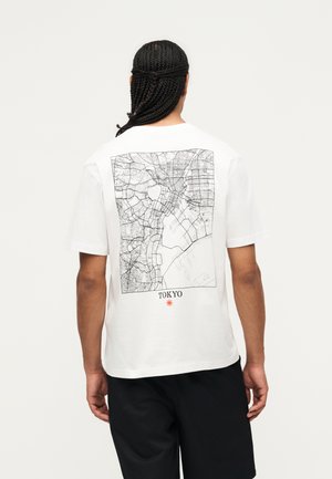 Person mit geflochtenem Haar trägt weißes Shirt, das auf dem Rücken über schwarzen Shorts eine detaillierte schwarze Linienkarte von Tokio zeigt.