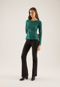 Green long-sleeve top with a round neckline, paired with black flared jeans and black ankle boots, standing against a neutral background.