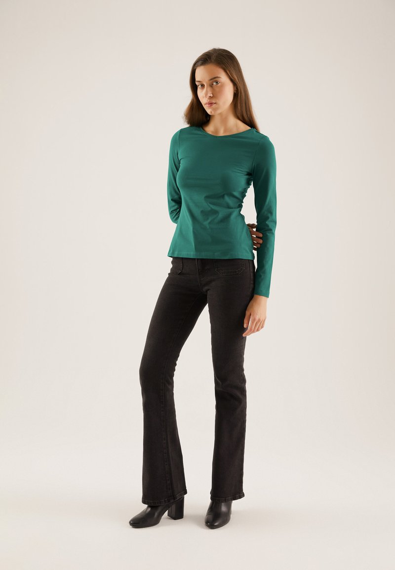 Green long-sleeve top with a round neckline, paired with black flared jeans and black ankle boots, standing against a neutral background.