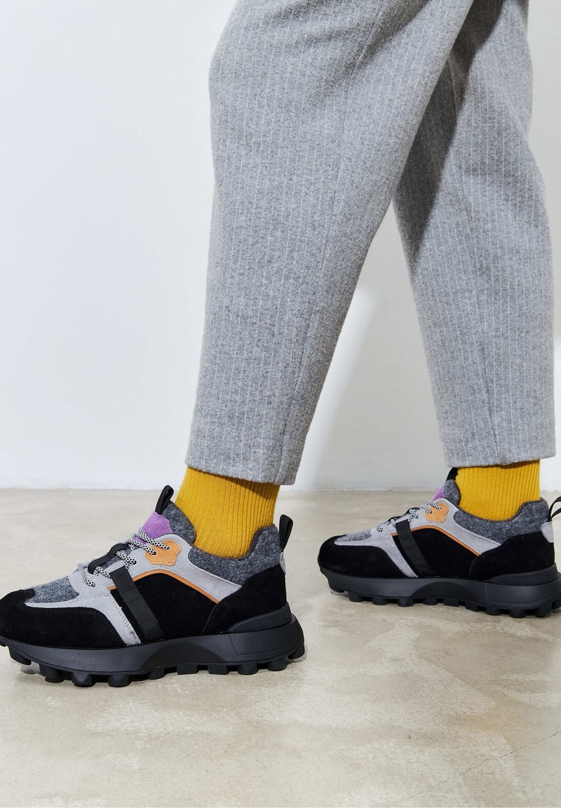 Personne portant un pantalon gris, des chaussettes jaunes et des baskets épaisses noires, grises et violettes, debout sur un sol lisse.