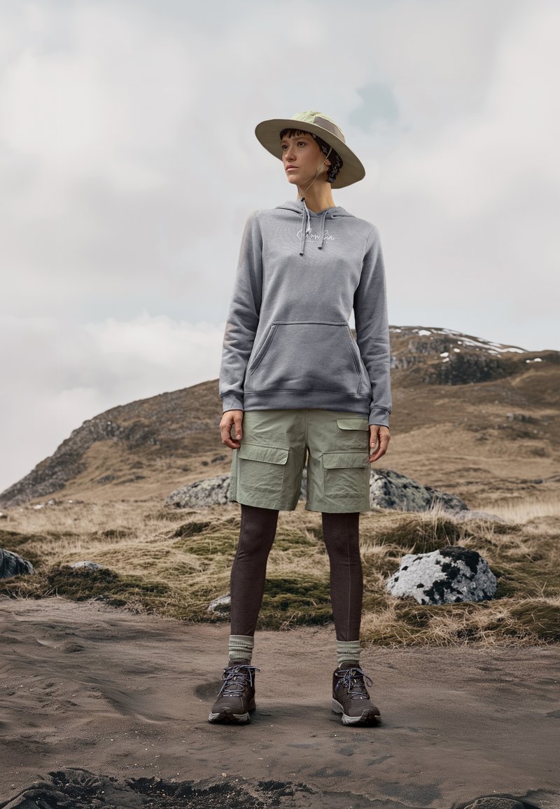 Sudadera gris claro con un bolsillo tipo canguro, pantalones cortos de cargo verdes, leggings marrones, gorra gris y verde, y zapatos de senderismo marrones en terreno rocoso.