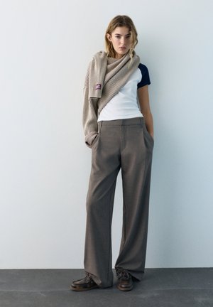 Fiatal nő bézs sállal, sötétkék-fehér pólóval, szürke bő szárú nadrággal és barna cipővel, egyszínű világos fal előtt állva.