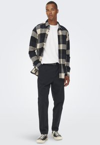 Camicia di flanella a quadri blu e grigia, indossata sopra una maglietta bianca, abbinata a pantaloni neri e sneakers nere con suole bianche.