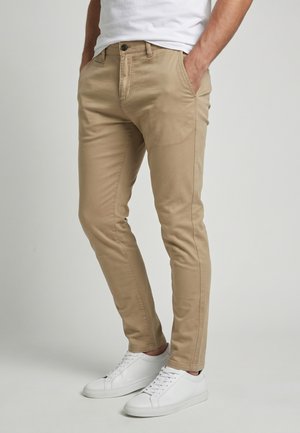 Homme portant un chino beige slim-fit avec les mains dans les poches et des baskets blanches basses sur un fond uni.