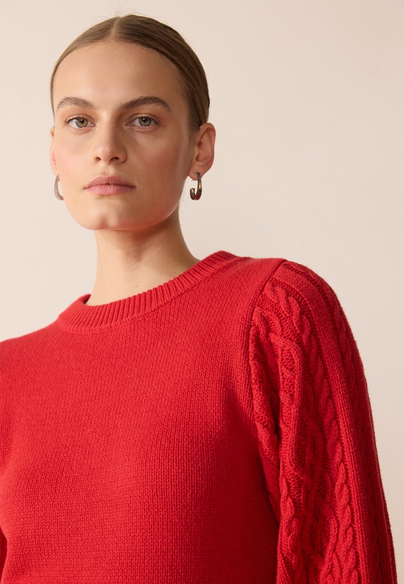 Czerwony sweter dzianinowy z prążkowanym okrągłym dekoltem, teksturowanym warkoczowym wzorem na rękawach oraz gładkim materiałem na korpusie.