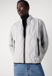 Quilted bomber jacket in light gray, featuring a textured wave pattern, zippered pockets, and a black zipper. Worn over a black turtleneck.