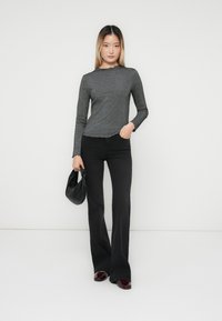 Donna con una maglietta grigia a maniche lunghe, pantaloni a zampa neri, scarpe bordeaux, che tiene una piccola borsa nera, in piedi contro uno sfondo bianco semplice.