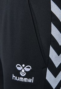 Czarne sportowe spodnie z białymi ukośnymi paskami i małym białym haftowanym logo. Wykonane z gładkiego, lekkiego materiału.