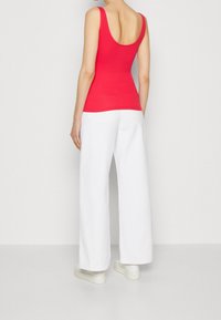 Person i en ärmlös röd topp och vida vita byxor, stående vänd bort från kameran mot en enfärgad vit bakgrund.