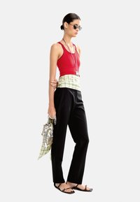 Czerwona, bezrękawnikowa bluzka, czarne spodnie i płaskie sandały. Nosi błyszczącą torbę ozdobioną cekinami oraz zieloną kraciastą chustę owiniętą wokół talii.