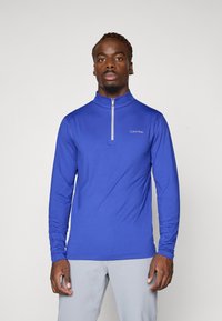Karališkai mėlynas ilgomis rankovėmis viršus su užtrauktuku ant apykaklės. Turi prigludusį dizainą ir baltą Calvin Klein logotipą kairiajame krūtinės kampe.