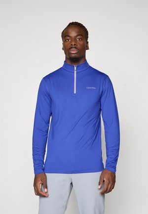 Royal blue bluzka z długim rękawem i zamkiem przy kołnierzu. Posiada dopasowany krój oraz białe logo Calvin Klein na lewej piersi.