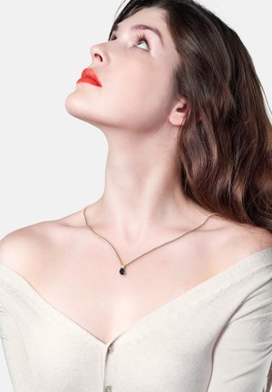 Goldene Halskette mit einem teardrop-förmigen schwarzen Edelstein-Anhänger. Das Model trägt ein beige Off-Shoulder-Oberteil. Glatte Haut, neutraler Hintergrund.