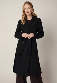 Czarny wełniany płaszcz trench z szerokim kołnierzem, paskiem w talii i bocznymi rozcięciami. Posiada duże guziki i strukturalne ramiona.