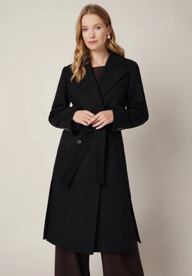 Czarny wełniany płaszcz trench z szerokim kołnierzem, paskiem w talii i bocznymi rozcięciami. Posiada duże guziki i strukturalne ramiona.