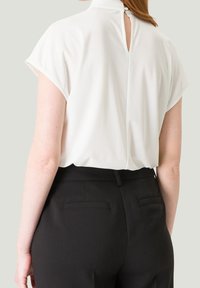 Witte blouse met korte mouwen, hoge halslijn en een keyhole-detail aan de achterkant, gecombineerd met zwarte tailored pantalons met achterzakken.