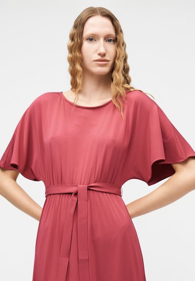 Γυναίκα με μακριά κυματιστά ξανθά μαλλιά φοράει ένα φαρδύ, κοντομάνικο, ρόζ φόρεμα με ζώνη, στέκεται με τα χέρια στη μέση μπροστά από ένα απλό φόντο.