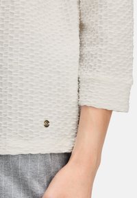 Haut blanc texturé à manches trois-quarts avec un petit détail de bouton doré près de l'ourlet, présenté avec un pantalon gris à rayures.