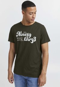 Olivengrønn bomulls t-skjorte med hvit grafisk tekst "Ninetgthr33" og "Blend Denim" på fronten. Korte ermer og rund hals.