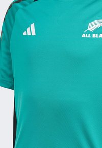 Türkise Sportshirt mit schwarzen Seitenpartien. Verfügt über weiße Markenkennzeichnung mit einem Blättermuster und drei weißen Streifen auf der linken Schulter.