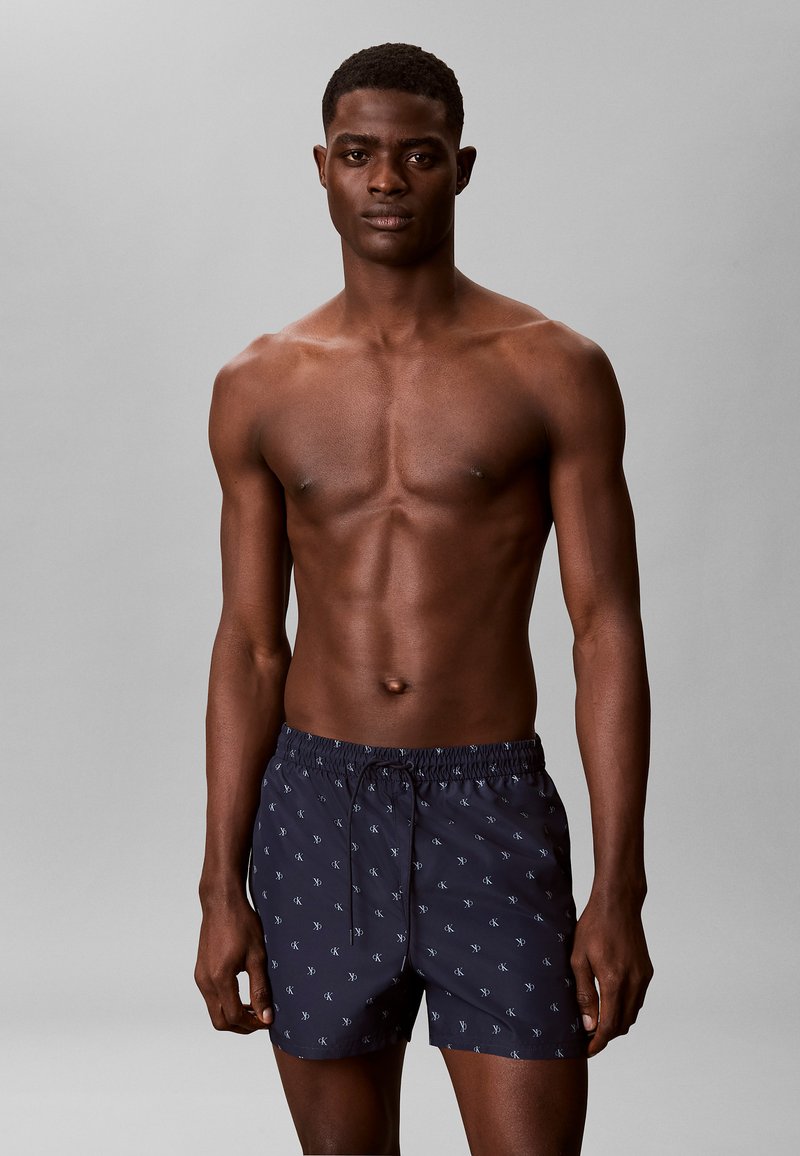 Homme torse nu portant un short de bain bleu marine à petit motif blanc, debout devant un fond gris uni.