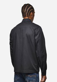 Schwarzes Baumwollhemd mit lässiger Passform, langen Ärmeln und Knopfmanschetten. Der Rücken verfügt über einen Schulterbesatz und dezente Nähte für Struktur.