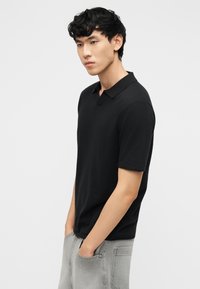 Νέος άνδρας με μαύρα σγουρά μαλλιά που φοράει μαύρο κοντομάνικο πόλο και ανοιχτό γκρι τζιν, με τα χέρια στις τσέπες, στραμμένος προς τα αριστερά.