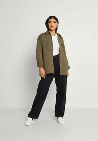 Mulher em pé, vestindo um conjunto casual com uma camisa verde-oliva abotoada, um crop top branco, calças largas pretas e ténis brancos, contra um fundo simples.