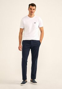 Witte katoen T-shirt met een zak, gecombineerd met marineblauwe broek. Model draagt marineblauwe schoenen en staat tegen een lichte achtergrond.
