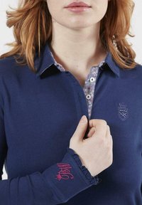 Polo shirt bleu marine avec un col à motif floral, comportant un logo en écusson sur la poitrine et un poignet froncé avec un détail de broderie rose.