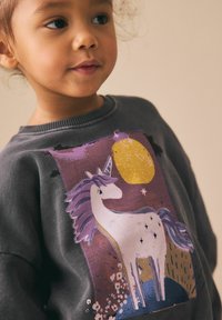 Niño pequeño con sudadera oscura que tiene una ilustración de un unicornio en púrpura y blanco, con una gran luna amarilla al fondo.