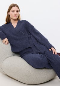 Maglione avvolgente lavorato a maglia di colore blu navy con maniche lunghe e pantaloni coordinati, indossato su una sedia curva con superficie grigio chiaro e texture. 