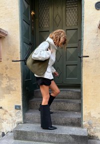 Bottes noires à talons hauts, ajustées à la cheville avec un bout pointu élégant. Sac tressé vert olive, veste blanche légère et shorts sombres. Marches en pierre.