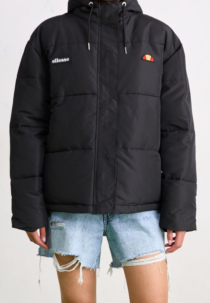 Doudoune noire à capuche, logo ellesse sur la poitrine, portée avec un short en jean bleu clair déchiré sur une personne, fond uni.