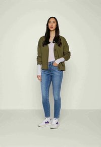 Cardigan verde-azeitona com botões pretos, blusa de manga longa cinza-claro, jeans skinny azuis e ténis brancos com detalhes roxos.