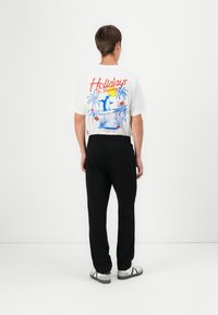 White cotton t-shirt with blue and red "Holidays in Paradise" graphic; paired with black pants and gray sneakers; casual fit.