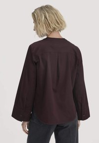 Camicia a maniche lunghe di colore bordeaux con taglio dritto, piega sul retro e dettagli dei polsini abbottonati. Realizzata in tessuto leggero con una texture liscia.