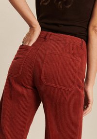 Pantalon en velours côtelé d'un rouge profond avec une texture à côtes verticales, deux poches arrière et une taille haute.