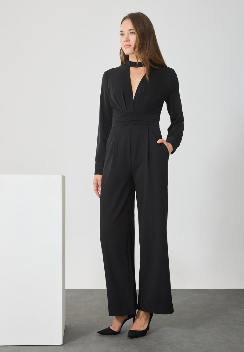 Combinaison noire avec un décolleté en V profond, des manches longues et une taille cintrée. Fabriquée en tissu lisse avec des jambes larges et des poches latérales.