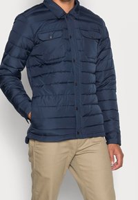Chaqueta acolchada de color marino con cierre de botones, dos bolsillos en el pecho y textura acolchada. Presenta un cuello con solapa y un diseño estratificado.