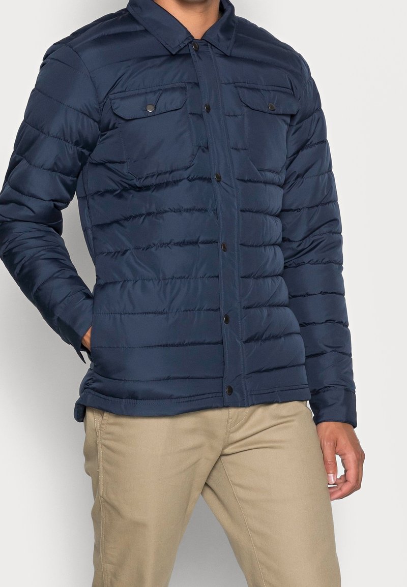 Chaqueta acolchada de color marino con cierre de botones, dos bolsillos en el pecho y textura acolchada. Presenta un cuello con solapa y un diseño estratificado.