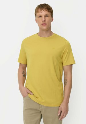 Ung mand med kort hår iført en gul t-shirt og beige bukser, står med den ene hånd i lommen mod en ensfarvet baggrund.