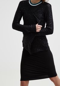 Svart stickad cardigan med en glittrande textur, långa ärmar och en crossover-design, i kombination med en figurnära svart kjol.