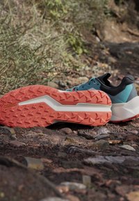 Trailrunning-Schuhe mit orangefarbenen Profilsohlen, die auf felsigem, natürlichem Untergrund mit grüner Vegetation liegen.