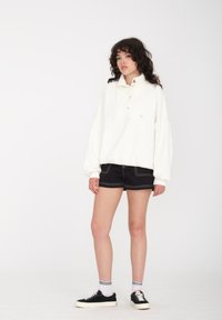 Witte oversized pullover met hoge kraag, voorzien van knopen, gedragen over zwarte shorts met witte stiksels, en zwarte sneakers met witte zolen.