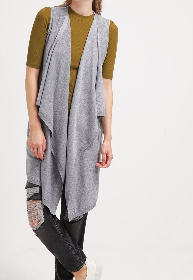 Cardigan gris sin mangas con un diseño drapeado, usado sobre una blusa de canalé verde oliva, combinado con jeans negros desteñidos y zapatos blancos.