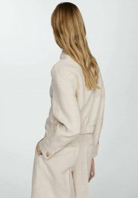 Beige linned blazer med en struktureret krave, elastisk talje og knapmanchetter, parret med matchende lyse bukser. Glat tekstur.