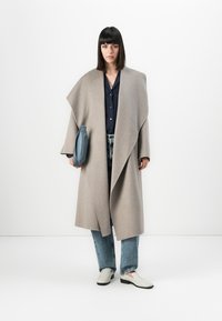 Ženska s ravnimi črnimi lasmi stoji oblečena v bež oversize plašč, mornarsko modro srajco, modre kavbojke, bele čevlje in drži modro torbico.